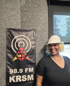 1000007848 A smiling woman wearing a white hat, silver hoop earrings, layered silver necklaces, and a black V-neck shirt stands inside a radio studio next to a black KRSM 98.9 FM banner. The banner features the station’s logo in red, orange, and yellow geometric shapes beneath concentric white circles. Gray acoustic wall panels and a window with outdoor light are visible in the background.